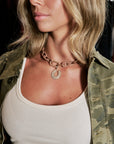 A close-up lifestyle shot of a woman wearing a chunky bronze chain necklace with a crescent moon pendant. She is wearing a white ribbed tank top and an open camouflage jacket.