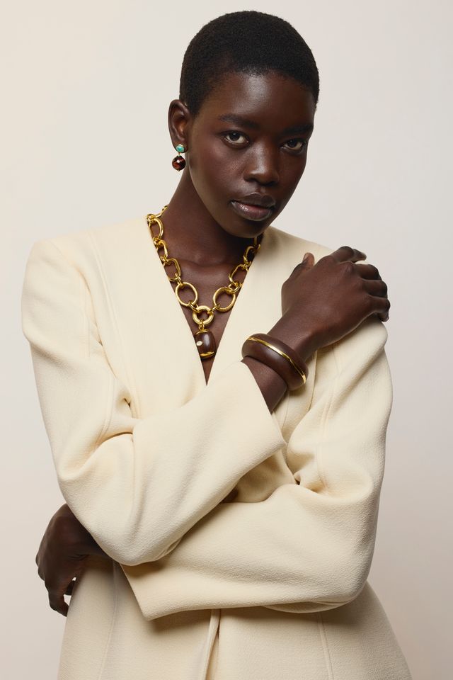 A model with short dark hair is wearing a cream-colored blazer, layered gold chain necklace, and gold bangles, Umbria Earrings in Tigers Eye set against a plain light beige background.