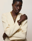 A model with short dark hair is wearing a cream-colored blazer, layered gold chain necklace, and gold bangles, Umbria Earrings in Tigers Eye set against a plain light beige background.