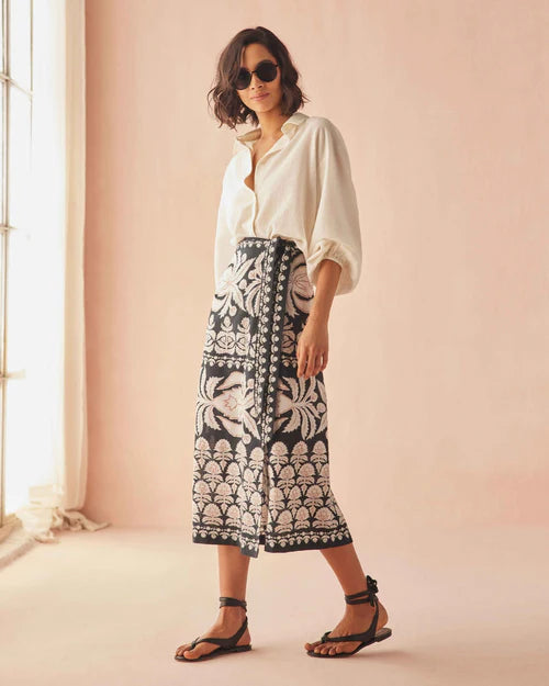 Woman wearing a white blouse and black patterned skirt against a pink wall. front/side
