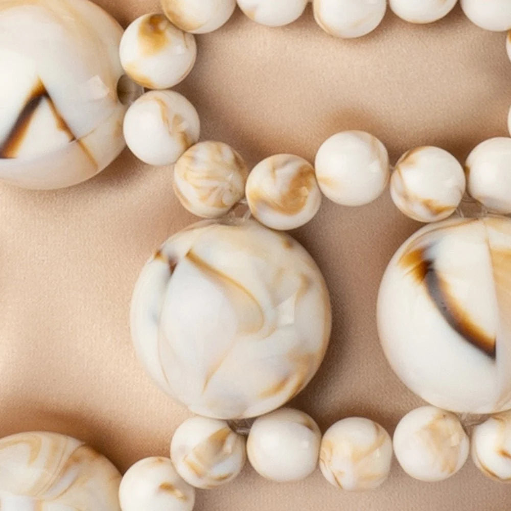 A close-up of a beaded necklace with large and small round beads in an off-white marbled pattern with tan and dark brown swirls, laid on a beige fabric background.