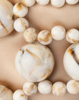 A close-up of a beaded necklace with large and small round beads in an off-white marbled pattern with tan and dark brown swirls, laid on a beige fabric background.