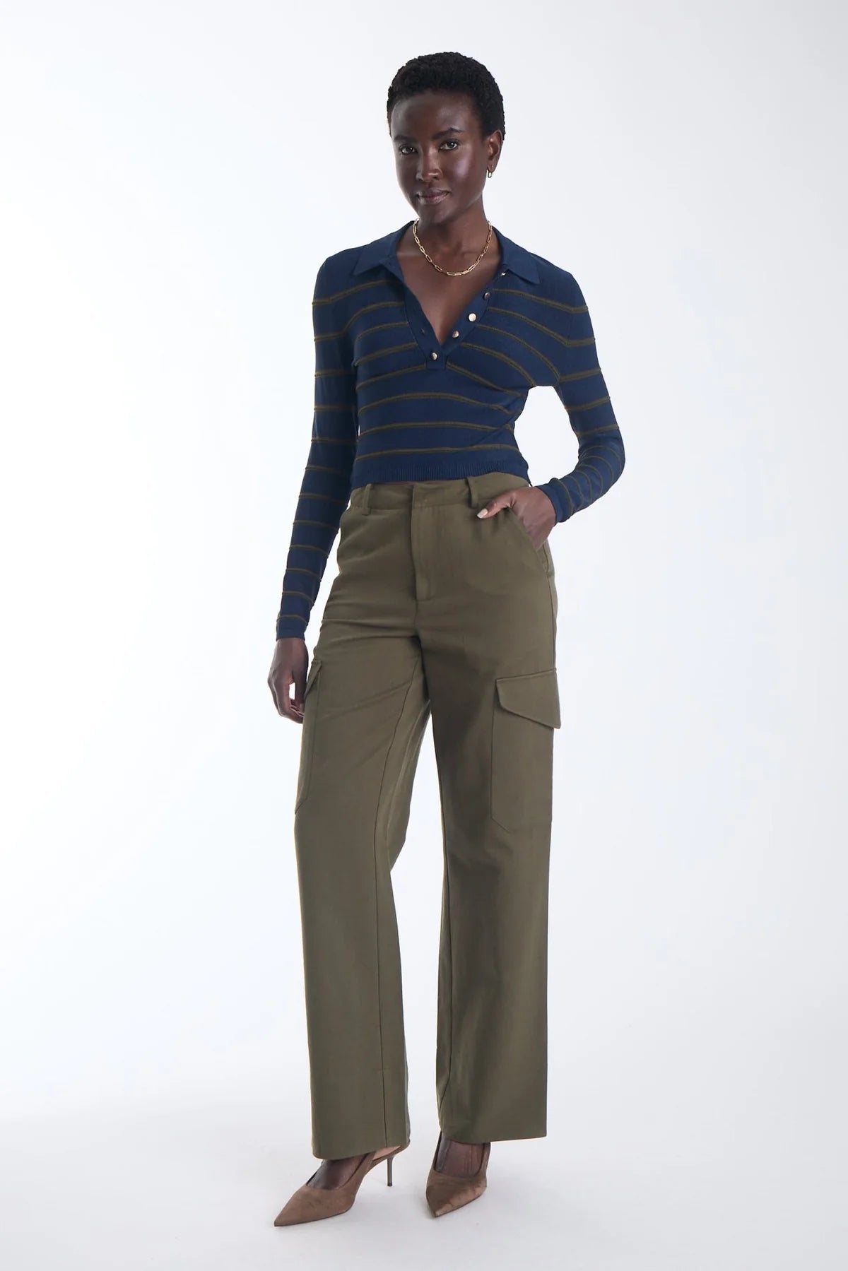 A model stands against a plain white studio background wearing a navy and gold striped long-sleeve polo shirt, olive green wide-leg cargo pants, and brown pointed-toe heels.