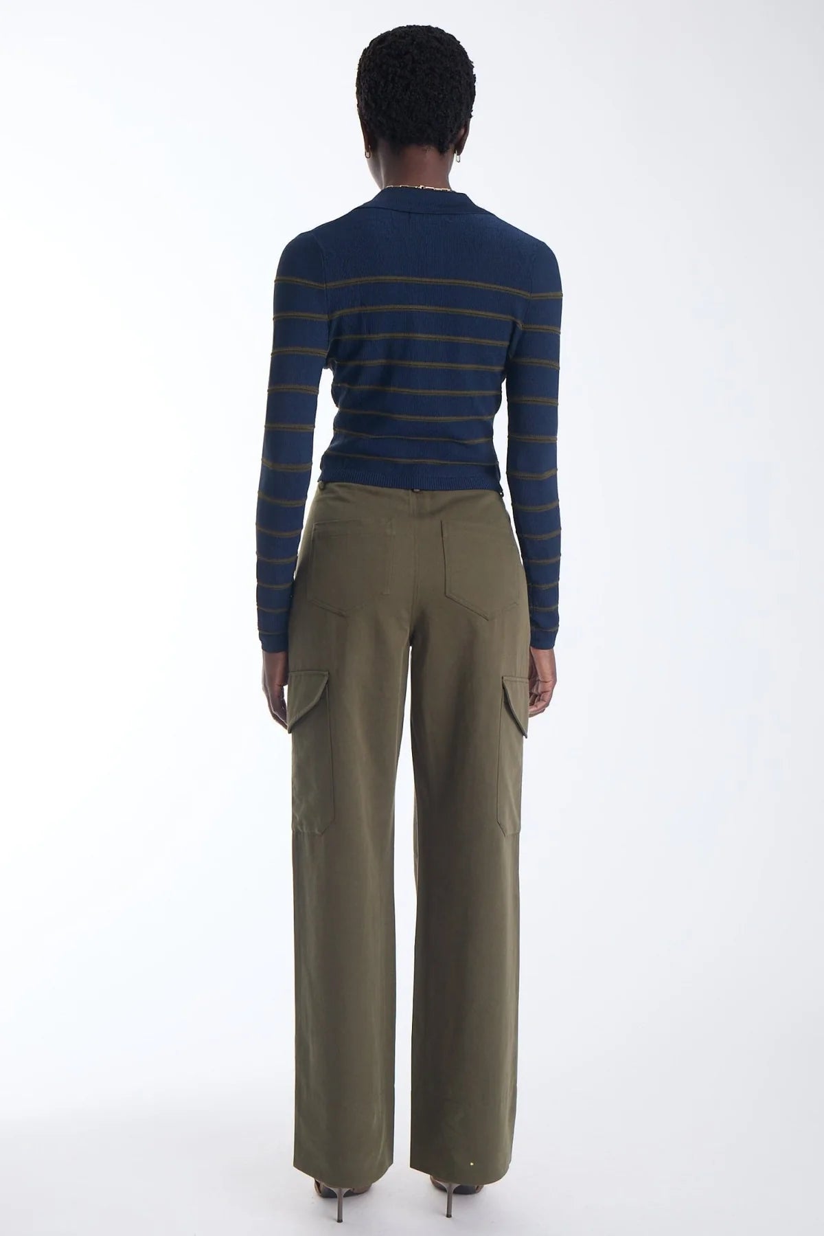A model shown from the back wearing a navy blue long-sleeve knit top with thin mustard yellow stripes and olive green wide-leg cargo pants, set against a plain white studio background.