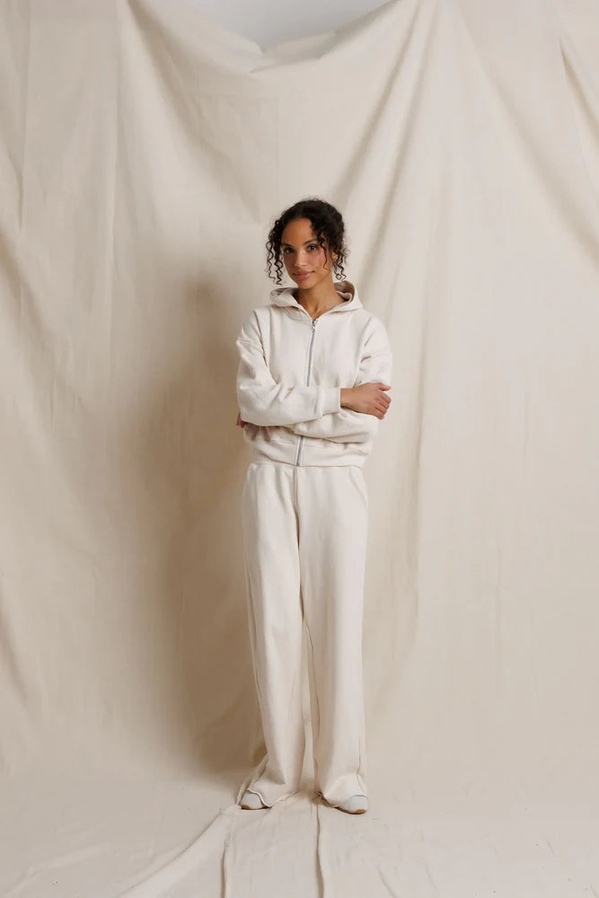 A model stands with arms crossed wearing a matching cream-colored zip-up hoodie and wide-leg sweatpants set, posed against a draped cream fabric backdrop.