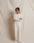 A model stands with arms crossed wearing a matching cream-colored zip-up hoodie and wide-leg sweatpants set, posed against a draped cream fabric backdrop.