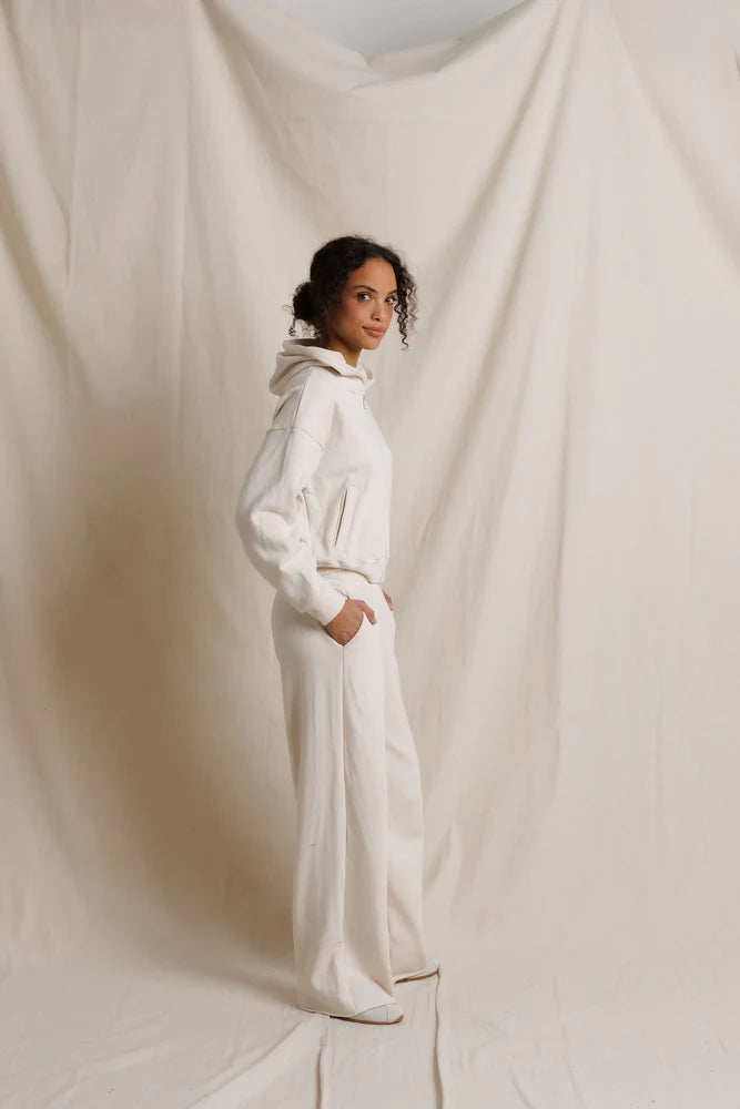 A woman wearing a matching white hooded sweatshirt and wide-leg sweatpants stands in profile against a draped cream fabric backdrop. side view