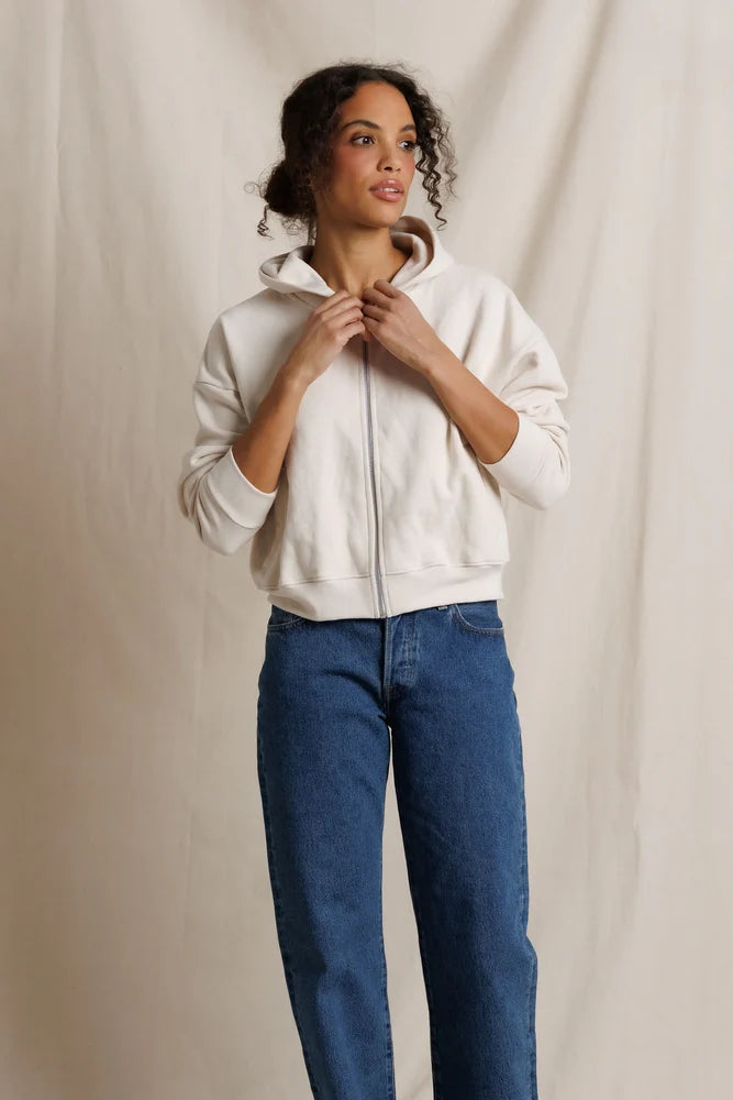 A woman wearing a cream-colored zip-up hoodie and blue jeans, standing against a neutral beige fabric backdrop.