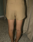 A person shown from the back wearing tan high-waisted shorts and a matching top, standing indoors on a speckled floor with curtains in the background.