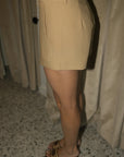 A person wearing a beige high-waisted mini skirt with pleated details and side pockets, paired with brown and beige heeled slide sandals, standing in front of beige curtains.