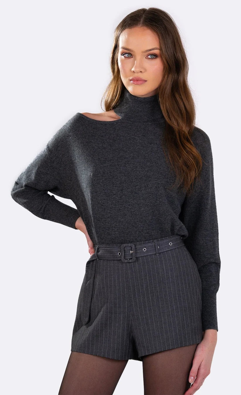 A woman modeling a dark gray knit turtleneck sweater with a shoulder cutout and matching dark gray pinstriped shorts with a belt, against a plain white studio background.