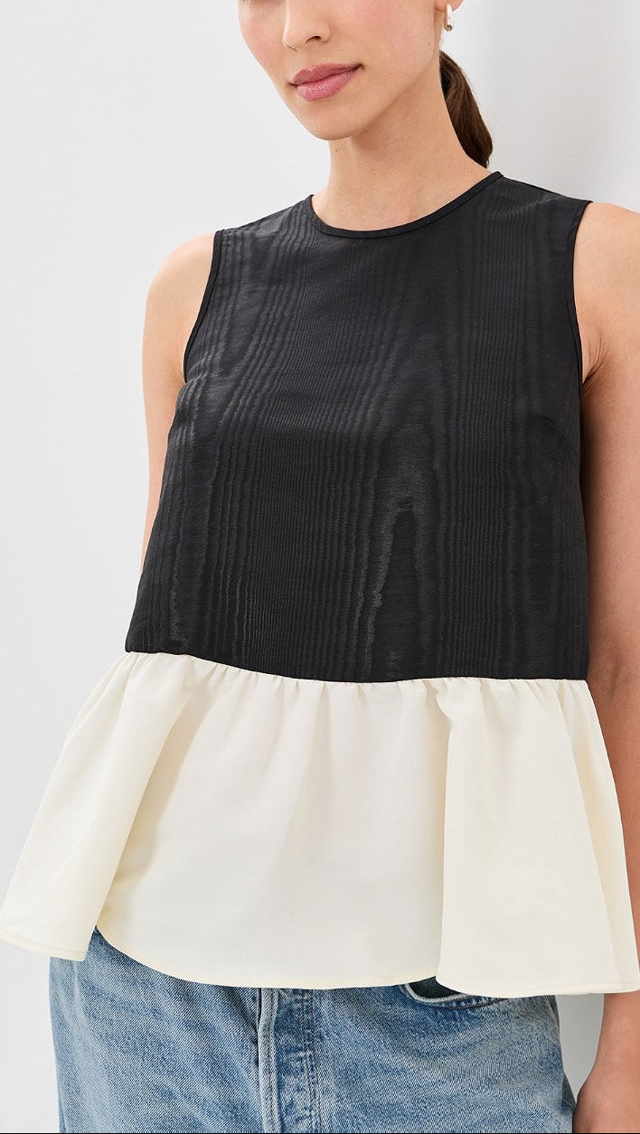 A woman wearing a sleeveless color-block top with a black textured upper section and a cream peplum lower section, paired with blue jeans against a light gray background.