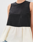 A woman wearing a sleeveless color-block top with a black textured upper section and a cream peplum lower section, paired with blue jeans against a light gray background.