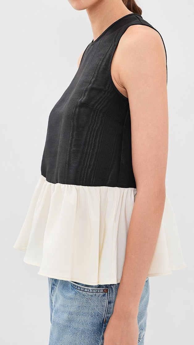 A woman modeling a sleeveless color-block top with a black upper section and an off-white flared lower section, paired with blue jeans against a light gray studio background.