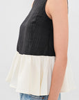 A woman modeling a sleeveless color-block top with a black upper section and an off-white flared lower section, paired with blue jeans against a light gray studio background.