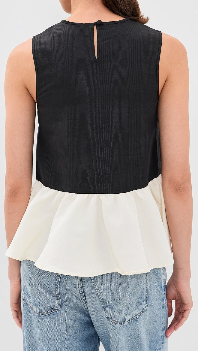 A woman shown from the back wearing a sleeveless color-block top with a black upper section and an off-white peplum lower section, paired with light blue jeans against a light gray studio background.