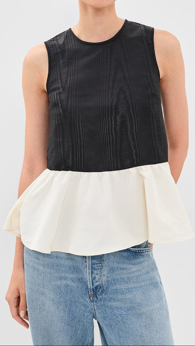 A woman modeling a sleeveless color-block top with a black textured upper section and an off-white peplum hem, paired with light blue jeans against a light gray studio background.