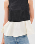 A woman modeling a sleeveless color-block top with a black textured upper section and an off-white peplum hem, paired with light blue jeans against a light gray studio background.