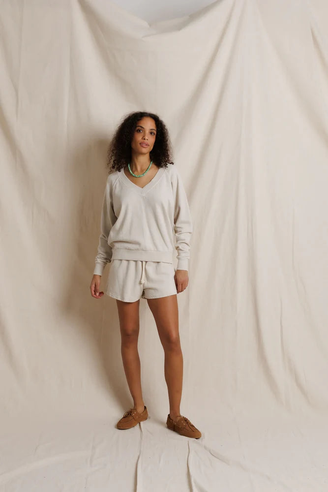 A woman stands against a draped cream fabric backdrop wearing a matching cream lounge set consisting of a long-sleeve V-neck top and drawstring shorts, paired with brown suede shoes and a green necklace.