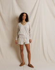 A woman stands against a draped cream fabric backdrop wearing a matching cream lounge set consisting of a long-sleeve V-neck top and drawstring shorts, paired with brown suede shoes and a green necklace.
