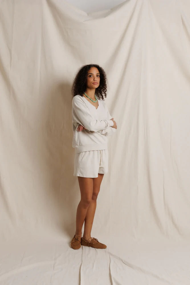 A woman stands in front of a draped cream fabric backdrop, wearing a matching off-white lounge set consisting of a long-sleeve top and shorts, paired with brown suede loafers.