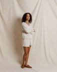 A woman stands in front of a draped cream fabric backdrop, wearing a matching off-white lounge set consisting of a long-sleeve top and shorts, paired with brown suede loafers.
