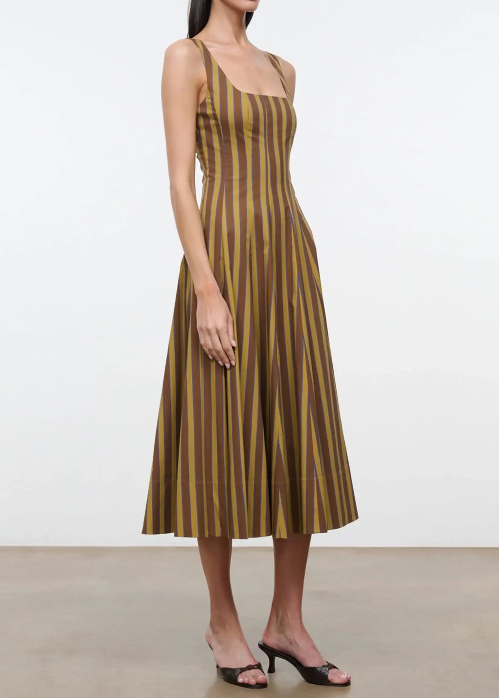 A woman modeling a sleeveless midi dress with vertical brown and olive-gold stripes, paired with black strappy heeled sandals, against a plain white studio background.