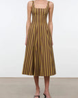 A woman modeling a sleeveless midi dress with vertical brown and mustard yellow stripes, standing against a plain white studio background.