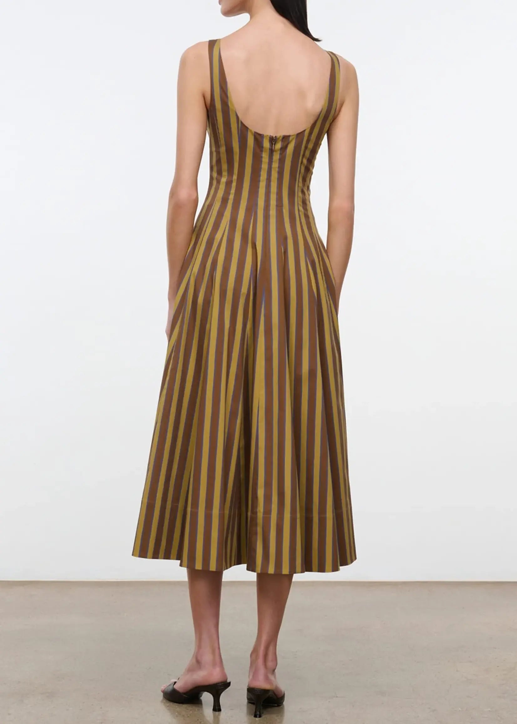 A woman shown from the back wearing a sleeveless midi dress with vertical brown and olive-gold stripes and a deep open back, paired with black heeled sandals against a plain studio background.