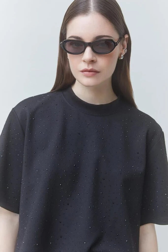 A woman wearing a black short-sleeve crewneck t-shirt with subtle silver rhinestone embellishments, styled with black cat-eye sunglasses against a light gray background.