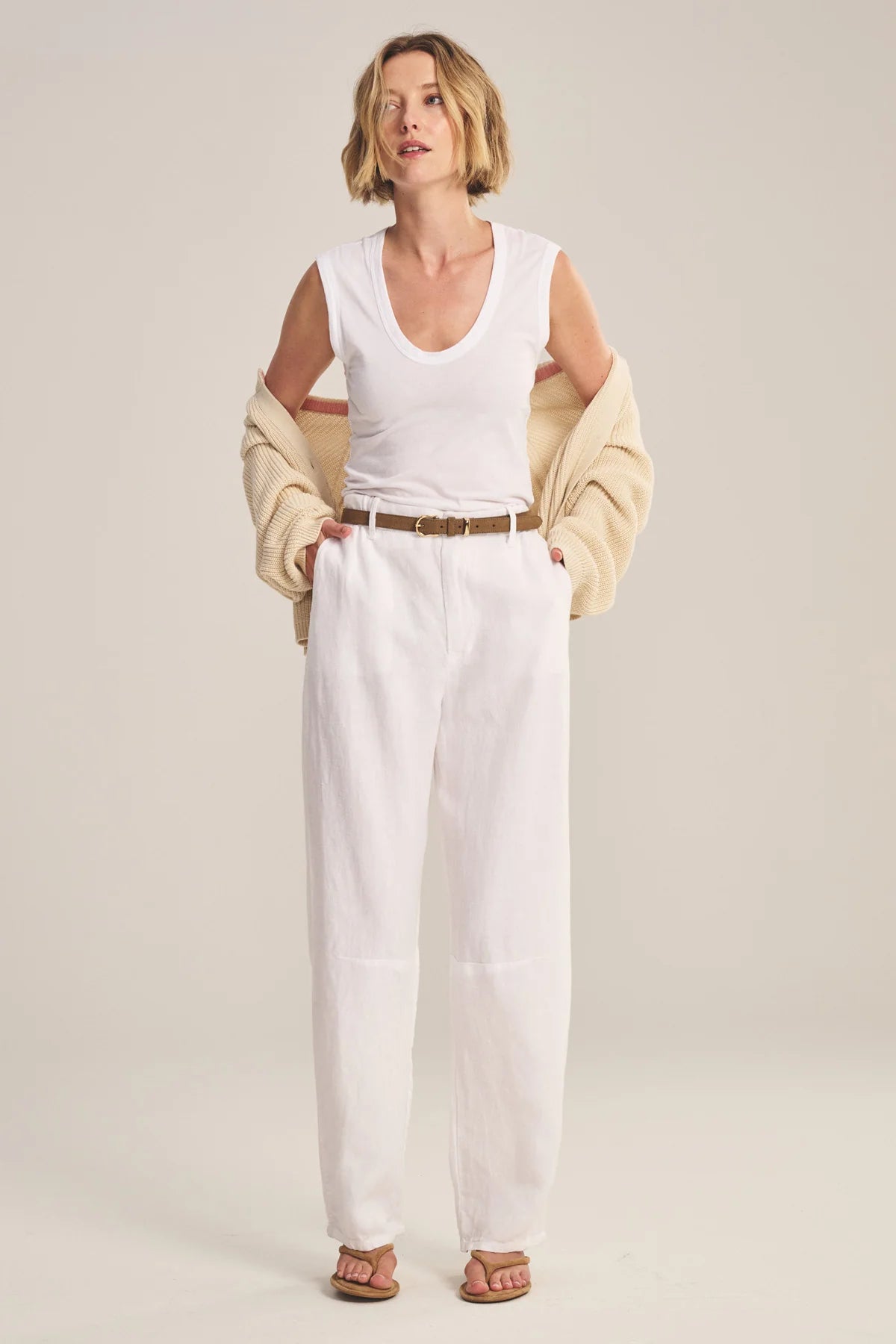 Woman wearing a white sleeveless top and pants with a beige cardigan on a plain background