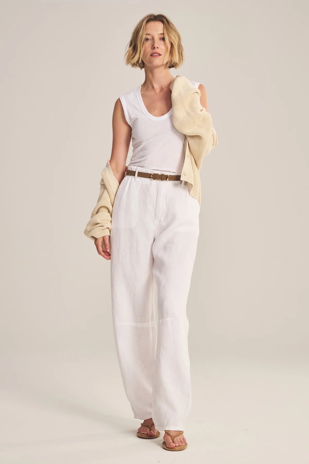 Woman wearing a white sleeveless top and pants with a beige jacket on a plain background