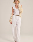 Woman wearing a white sleeveless top and pants with a beige jacket on a plain background