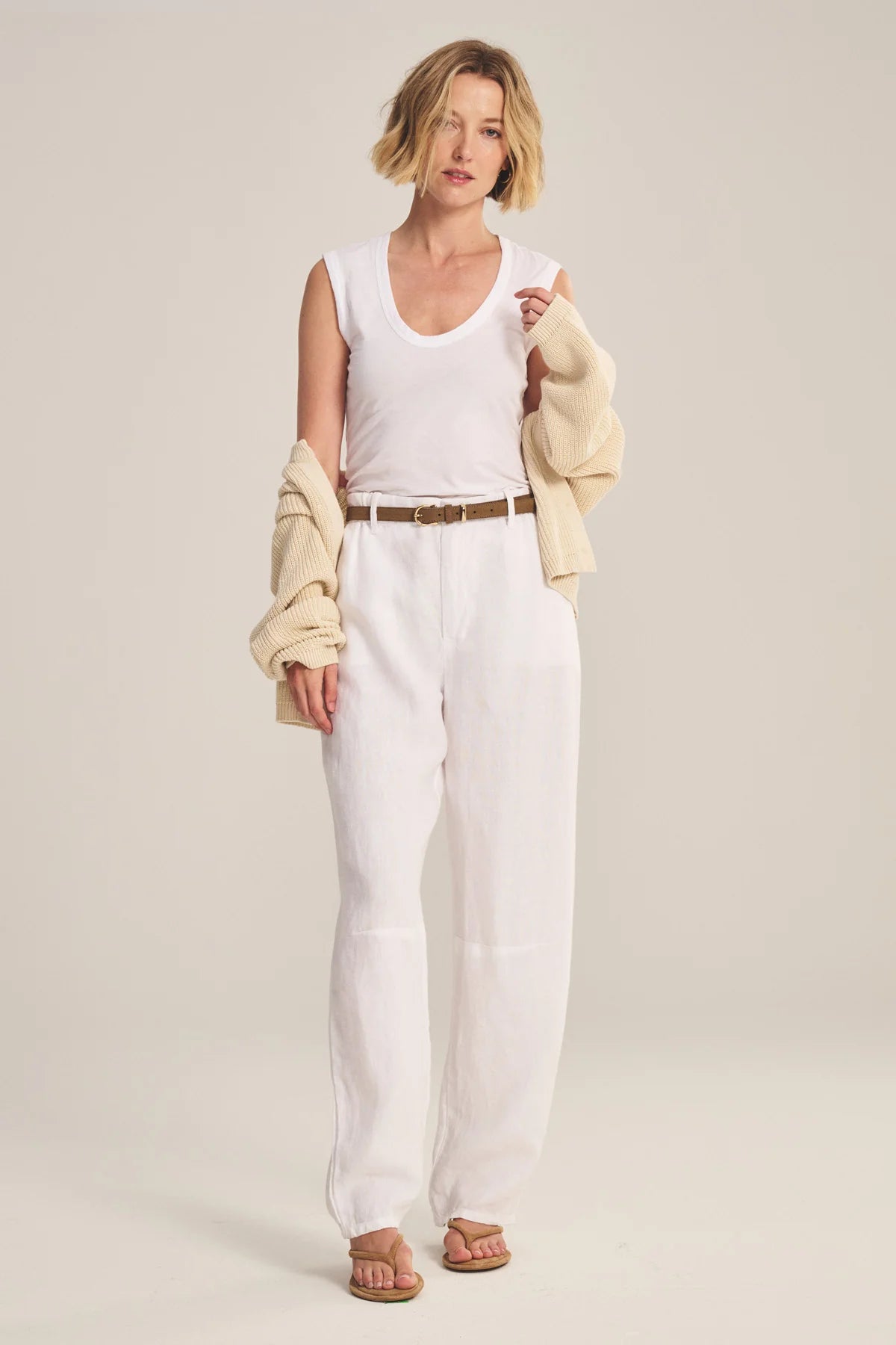 Woman wearing a white sleeveless top and pants with a beige cardigan on a plain background