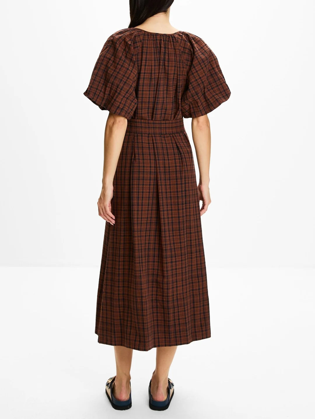 A woman viewed from the back wearing a brown and black plaid midi dress with puff sleeves and a tie waist, paired with black flat shoes, against a plain white studio background.