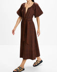 A woman modeling a brown and black plaid midi dress with puff sleeves, a deep V-neckline, and a matching waist tie, paired with black double-strap sandals against a plain white studio background.