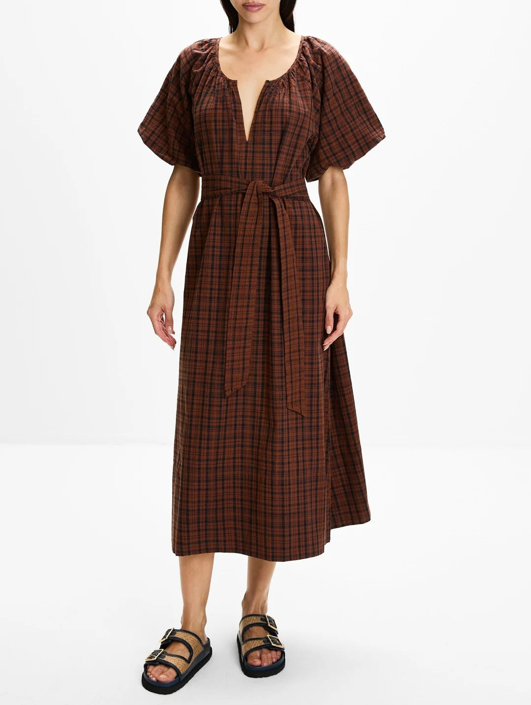 A woman modeling a brown and black plaid midi dress with puff sleeves, a deep V-neckline, and a matching fabric tie belt at the waist, paired with black strappy sandals against a plain white studio background.