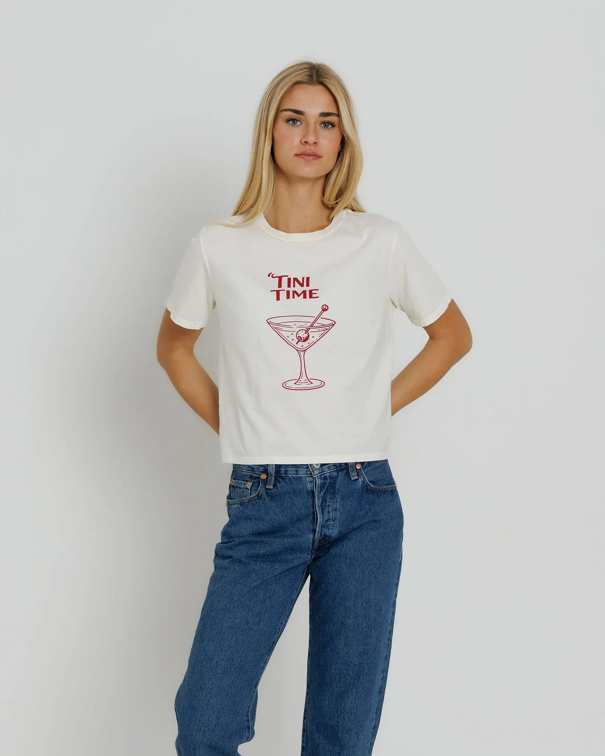 A woman wearing a white short-sleeve graphic t-shirt with a red 'TINI TIME' martini glass illustration on the front, paired with blue jeans, against a light gray studio background.
