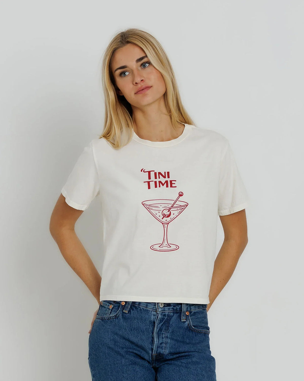 A woman wearing a white short-sleeve graphic t-shirt with a red 'TINI TIME' martini glass illustration on the front, paired with blue jeans, against a light gray studio background.