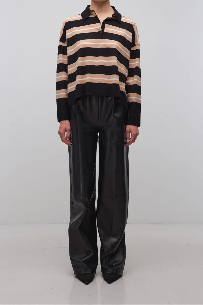 A model stands against a light gray studio background wearing a black and beige horizontally striped knit polo sweater with black wide-leg pants.