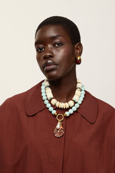 A model wearing Toro Earrings in Aubergine and a rust-brown oversized collared shirt and layered necklaces, including a prominent light blue and cream beaded necklace with a gold pendant, against a plain off-white background.