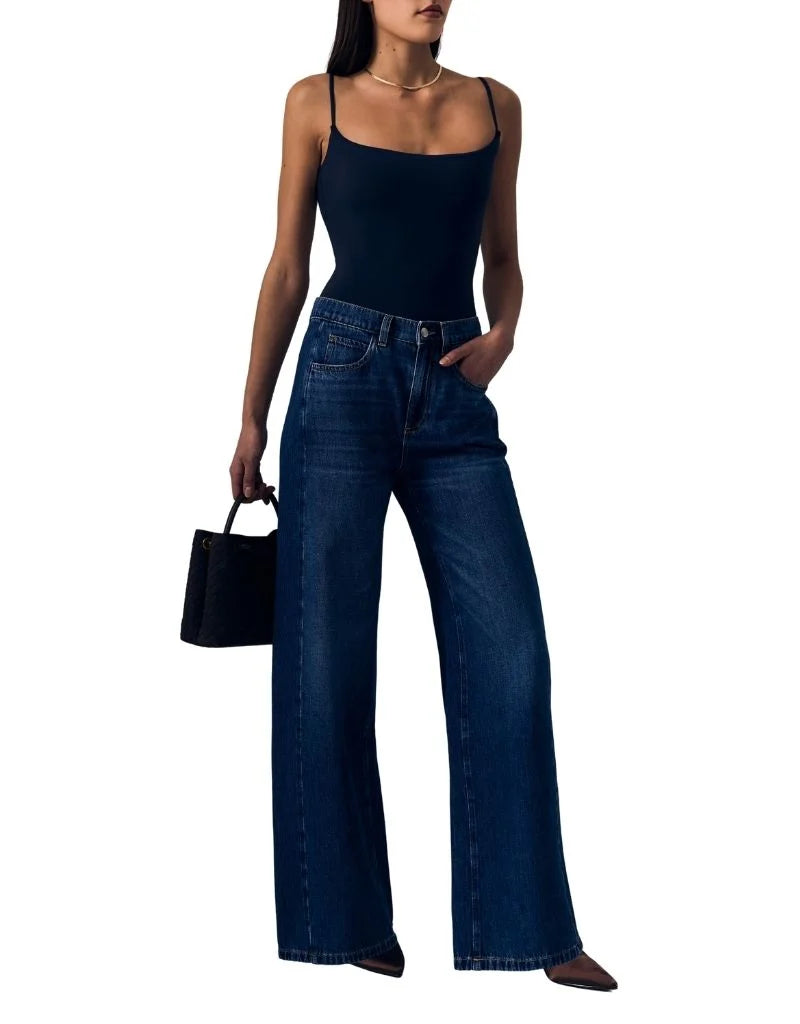 A woman modeling dark blue wide-leg jeans with a black camisole top, holding a small black handbag against a plain white background.