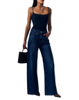 A woman modeling dark blue wide-leg jeans with a black camisole top, holding a small black handbag against a plain white background.