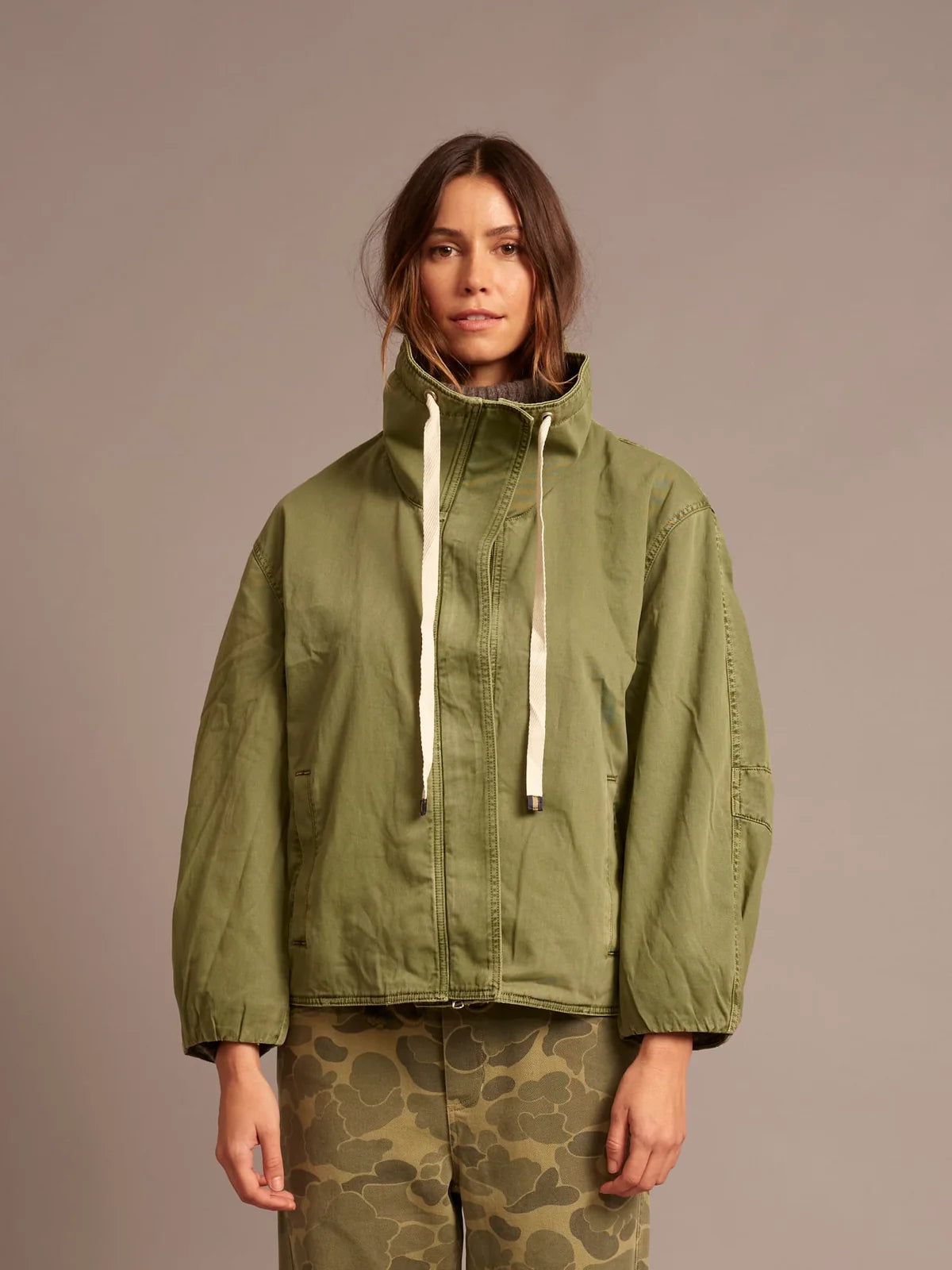 A woman wearing an olive green hooded jacket with cream drawstrings and olive camouflage pants, standing against a solid taupe background.