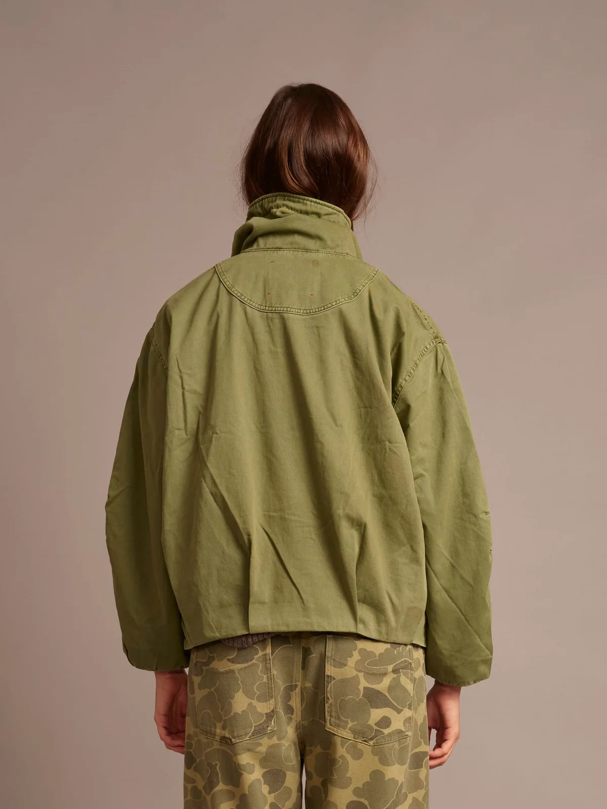 A person shown from the back wearing an olive green cropped jacket and olive camouflage pants against a plain warm beige studio background.