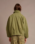A person shown from the back wearing an olive green cropped jacket and olive camouflage pants against a plain warm beige studio background.