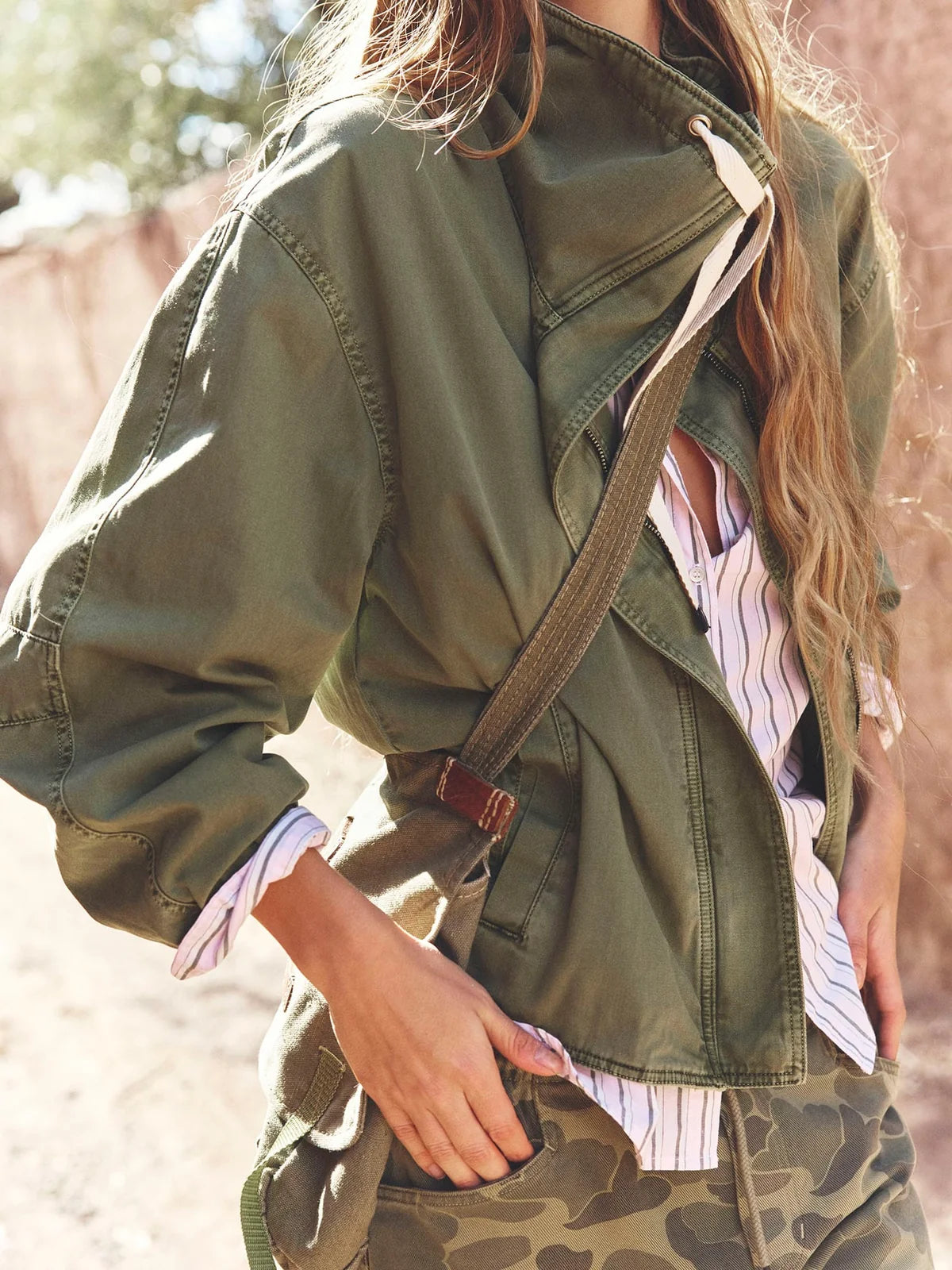 A woman wearing an olive green utility jacket over a white and navy striped shirt, paired with camouflage pants and a brown crossbody bag, in a warm outdoor setting.