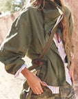 A woman wearing an olive green utility jacket over a white and navy striped shirt, paired with camouflage pants and a brown crossbody bag, in a warm outdoor setting.