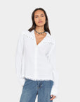 A woman modeling a white long-sleeve button-up shirt with silver stud embellishments on the shoulders and a frayed hem, paired with blue jeans against a light gray background.
