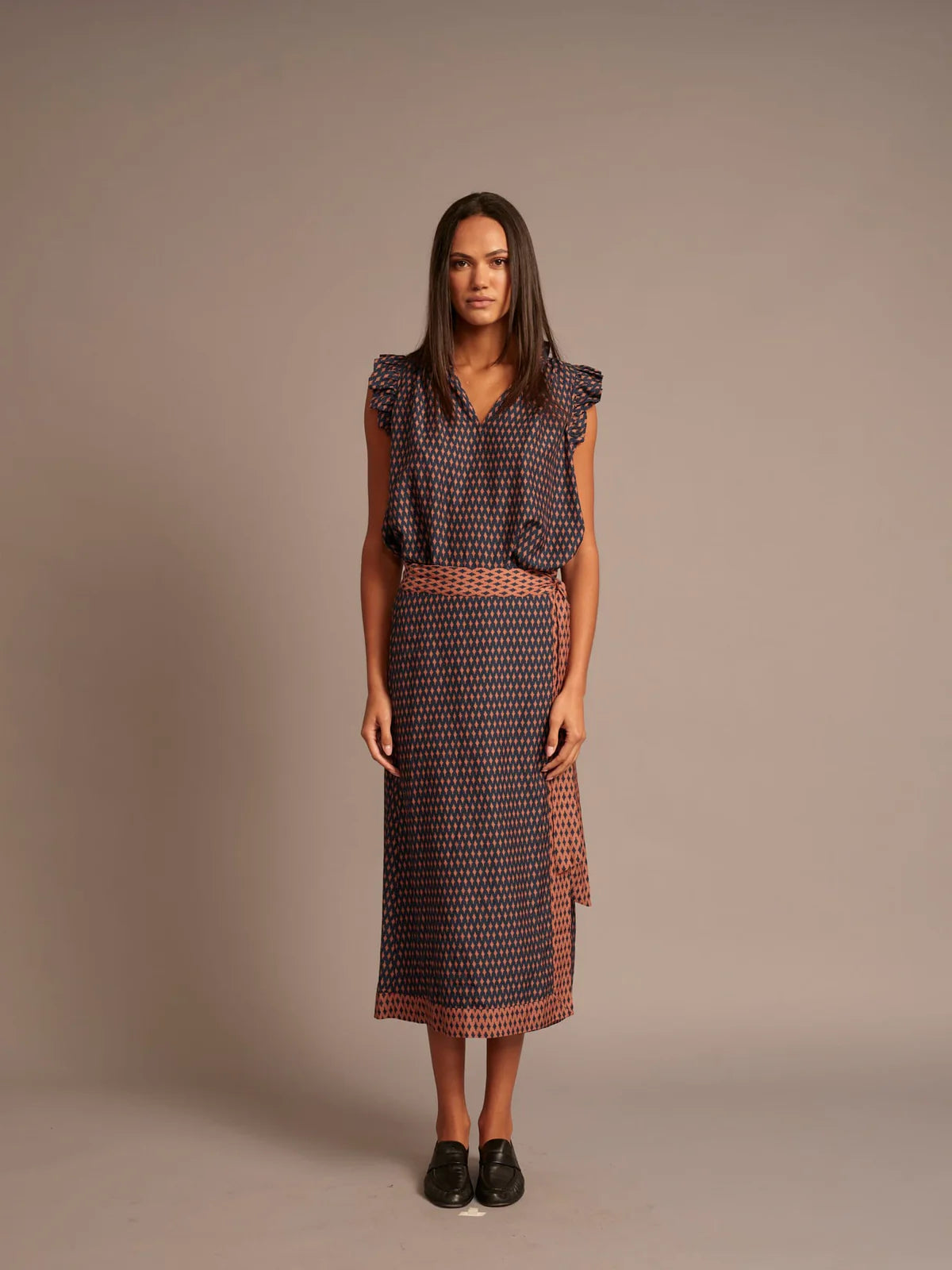 A woman stands against a plain taupe studio background wearing a sleeveless midi dress with a dark navy and rust red geometric pattern, ruffled cap sleeves, and a matching patterned belt.
